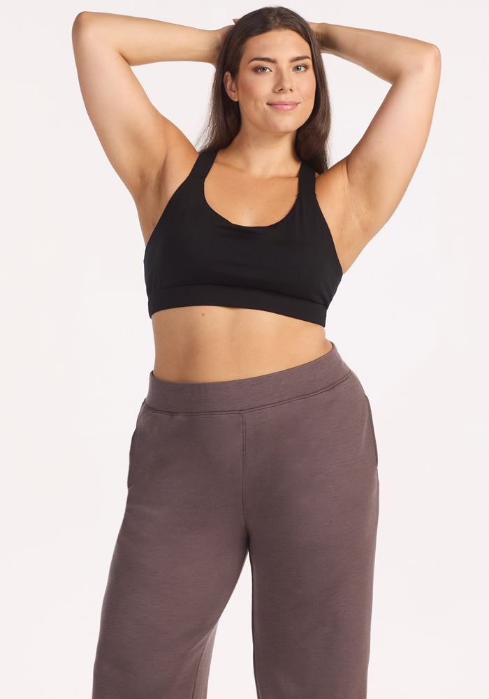 A woman with long brown hair poses confidently, wearing a black sports bra and Woolx Bree Lounge Pants in Plum Truffle, with her arms raised and hands behind her head against a plain white background.