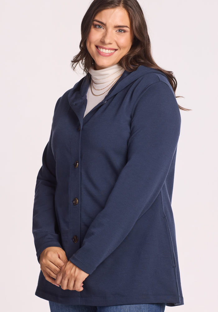 A woman with long brown hair smiles at the camera, wearing the Woolx Edna Hooded Cardigan in Deep Navy over a white turtleneck. She stands against a plain light background.