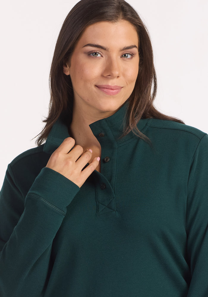 A woman with long brown hair and light skin smiles gently, wearing the Woolx Annie 3 Button Pullover in Deep Timber. She holds the collar with one hand and stands against a plain white background.