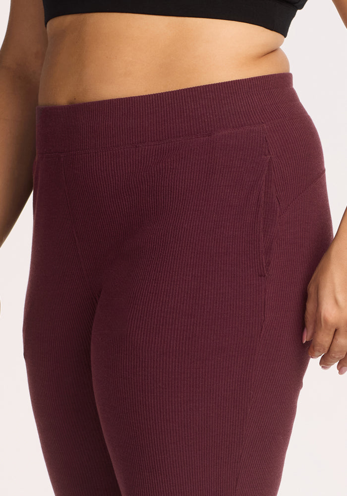 A person wearing Woolx Luca Ribbed Pants in Toasted Garnet, shown from waist to mid-thigh. The high-waisted pants feature a snug fit, ribbed texture, and visible seam details, paired with a black crop top.
