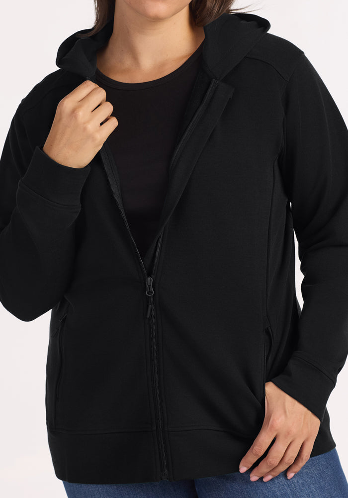 A person wears the Woolx Darby Zip Up Hoodie in black over a black T-shirt and blue jeans, holding the hoodie near the collar with one hand and resting the other near a pocket against a plain light background.