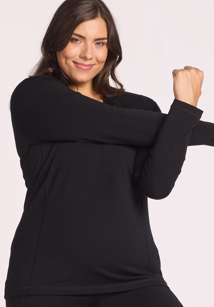 A woman with long brown hair, wearing the Woolx Elsa Long Sleeve in black, smiles confidently while stretching her right arm across her chest against a light background.