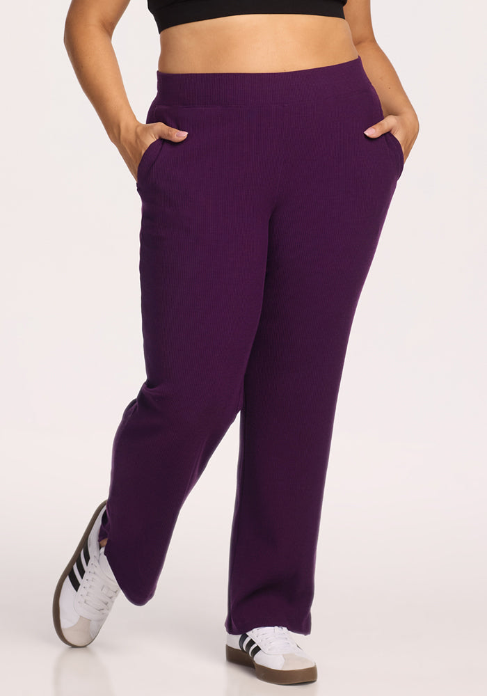 A person wears Woolx Luca Ribbed Pants in Majestic Purple, styled high-waisted with hands in pockets, paired with a black crop top and white sneakers with black stripes, standing against a plain background.