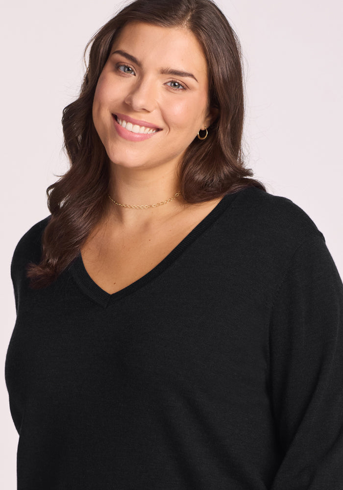 A woman with long brown hair, wearing the Woolx Francesca Sweater in black and a gold necklace, smiles softly against a light background.