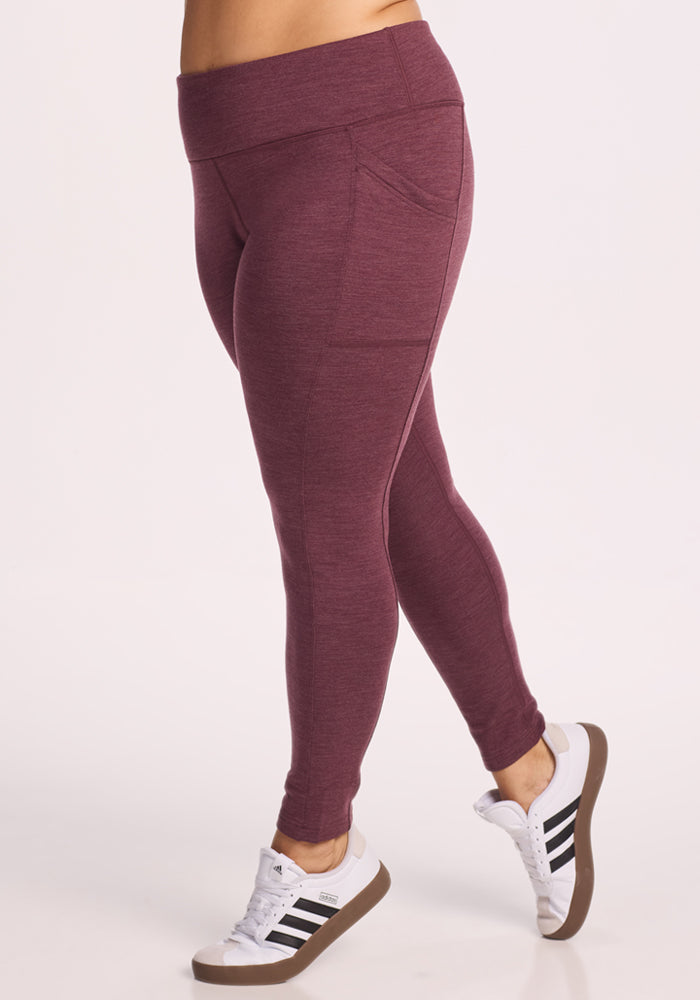 A person wearing Woolx Frost Duralite® Leggings in Grape Harvest Melange with side pockets and white Adidas sneakers with black stripes, standing against a plain light background.