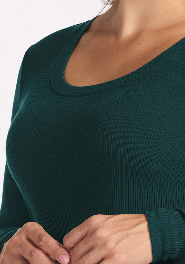 A close-up shows a person wearing the Woolx Caroline Ribbed Twirl Dress in Deep Timber. The ribbed merino dress features a scoop neckline, and the person's hands are gently clasped near their waist. Their head is not visible in the image.