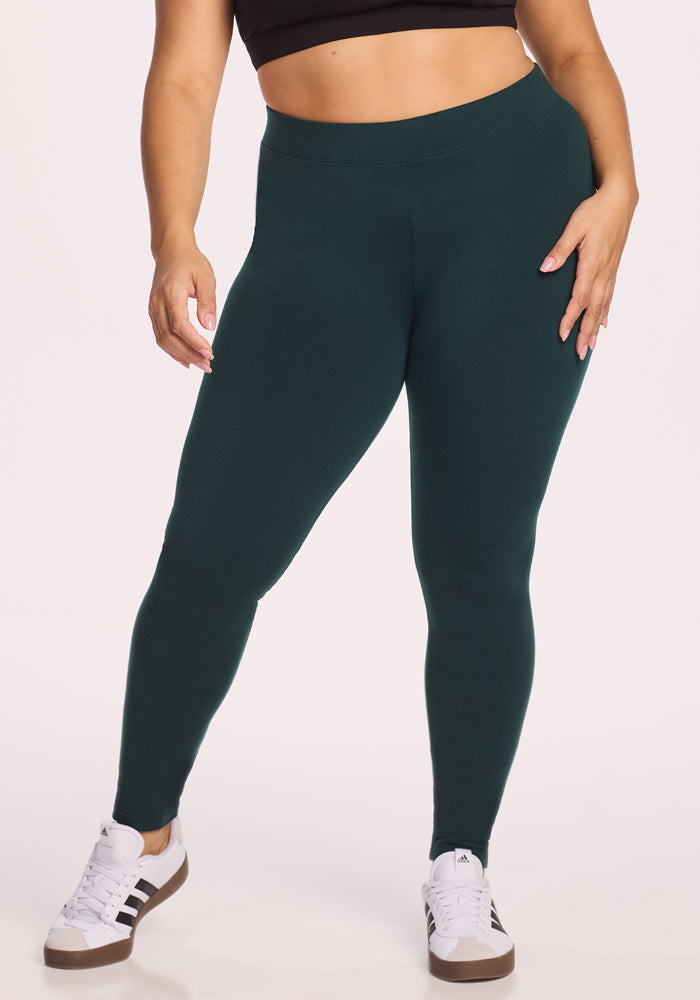 Against a plain light background, a person wears Woolx Stella Leggings in Deep Timber, a black crop top, and white Adidas sneakers with black stripes, resting their left hand on their hip. 