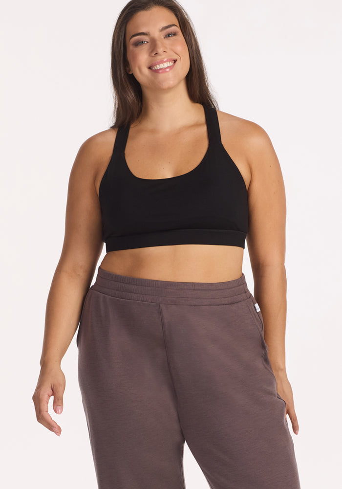 A woman with long brown hair smiles while wearing a black sports bra and Woolx Parker Sweatpants in Plum Truffle, standing against a plain light background.