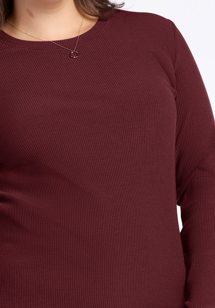 A person wears the Woolx Hadley Ribbed Crew in Toasted Garnet, showcasing its long sleeves and ribbed merino wool—ideal for lightweight travel layering. A delicate necklace with two interlocking circles complements the look. Only upper torso and part of head are visible.
