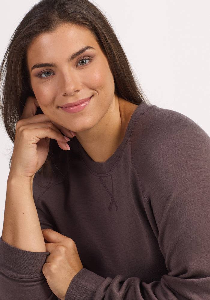 A woman with long brown hair, wearing the Woolx Bailey Crewneck Sweatshirt in Plum Truffle, featuring a recycled polyester exterior and soft Merino wool interior, smiles gently and rests her chin on her hand against a light background.