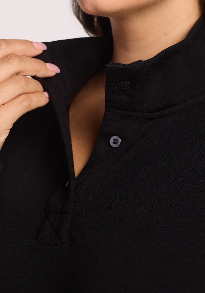 Close-up of a hand with light pink nails adjusting the collar and buttons of the Woolx Annie 3 Button Pullover in black. Only the lower face and neck are partially visible, with focus on the collar detail.