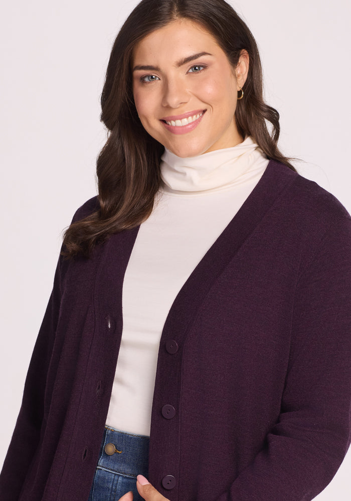A woman with long brown hair smiles while wearing the Woolx Gia Sweater in Deep Plum over a white turtleneck and blue jeans, standing against a plain light background.