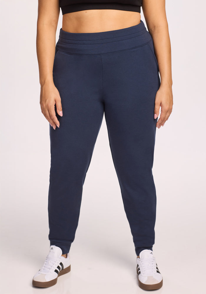 A person wearing Woolx Willa Joggers in Deep Navy, a black crop top, and white sneakers with black stripes stands against a plain light background. Only their body from shoulders to feet is visible. 