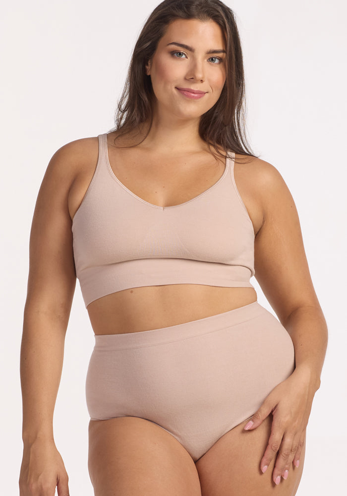 A woman with long brown hair smiles while wearing Woolx Mabel Full Coverage Underwear in Sand and a matching bralette, standing against a plain white background.