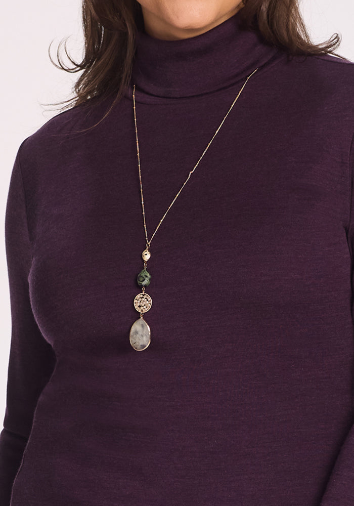 A person wears the Woolx Peyton Turtleneck in Deep Plum and a long gold necklace with three pendants: a round green bead, a filigree circle, and a large oval stone.