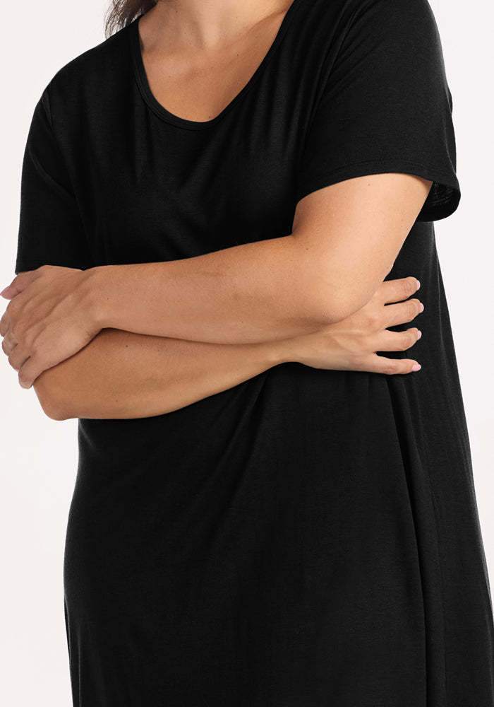 A person stands with arms crossed, wearing the Woolx Desi Nightgown in black—crafted from Merino wool for temperature-regulating comfort. The image is cropped from shoulders to mid-thigh.