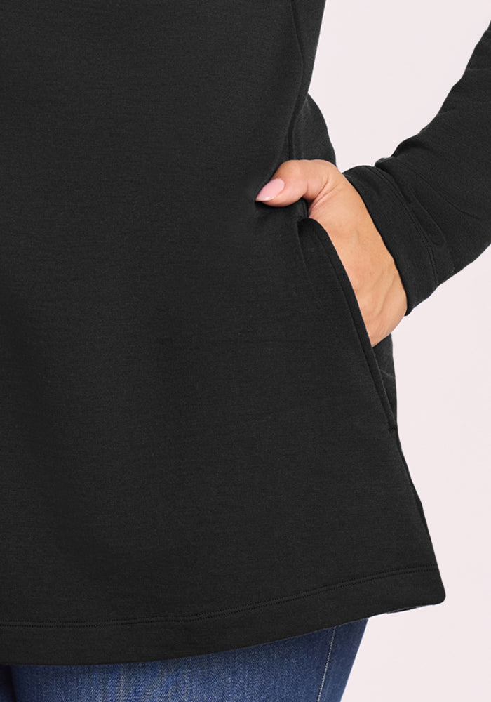 A person wearing the Woolx Edna Hooded Cardigan in black and blue jeans places their right hand in the cardigan's side pocket. The background is light-colored.