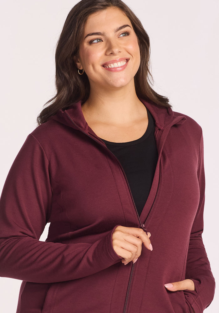 A smiling woman with long brown hair wears the Woolx Zoey Zip Up Hoodie in Toasted Garnet over a black top, standing against a light background.