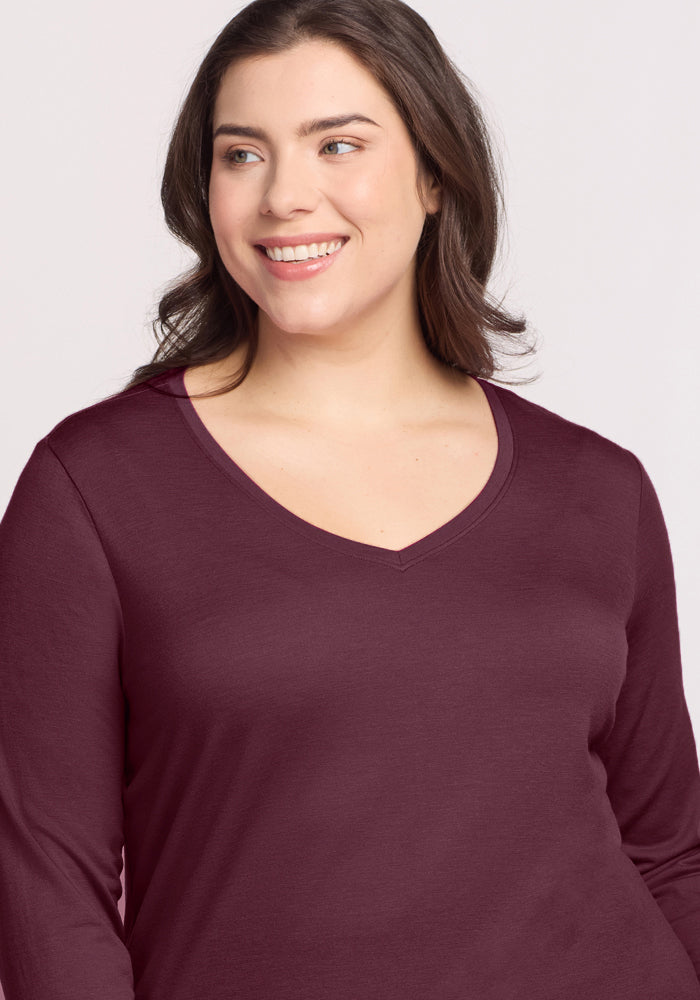 A woman with long brown hair smiles and looks to the side, wearing the Woolx Millie Long Sleeve in Grape Harvest, a Merino wool top known for comfort, against a plain light background.