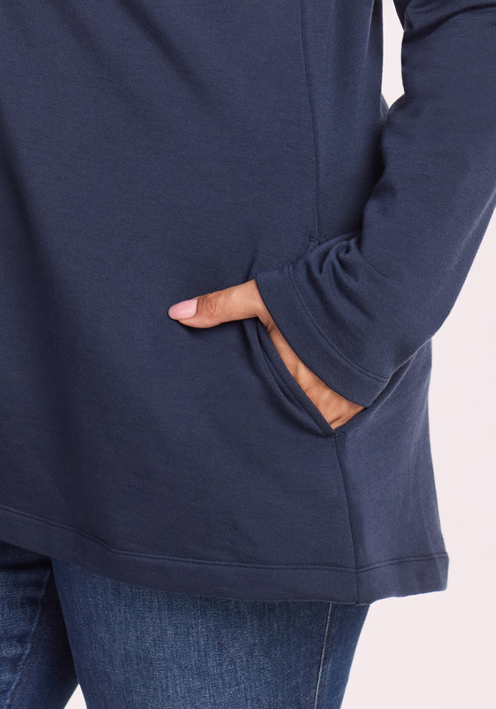 A person wears the Woolx Edna Hooded Cardigan in Deep Navy with blue jeans, hand in the pocket of the top. Only their lower torso, arm, and hand are shown, highlighting this cozy cold-weather style.