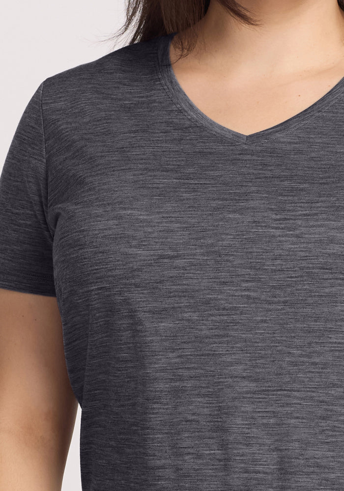 A close-up shows a person wearing the Woolx Junie V Neck in Cinder Melange—a dark gray, short-sleeve Merino wool tee. Only the neck, shoulder, and upper torso are visible; moisture-wicking fabric ensures comfort. The face is not shown.