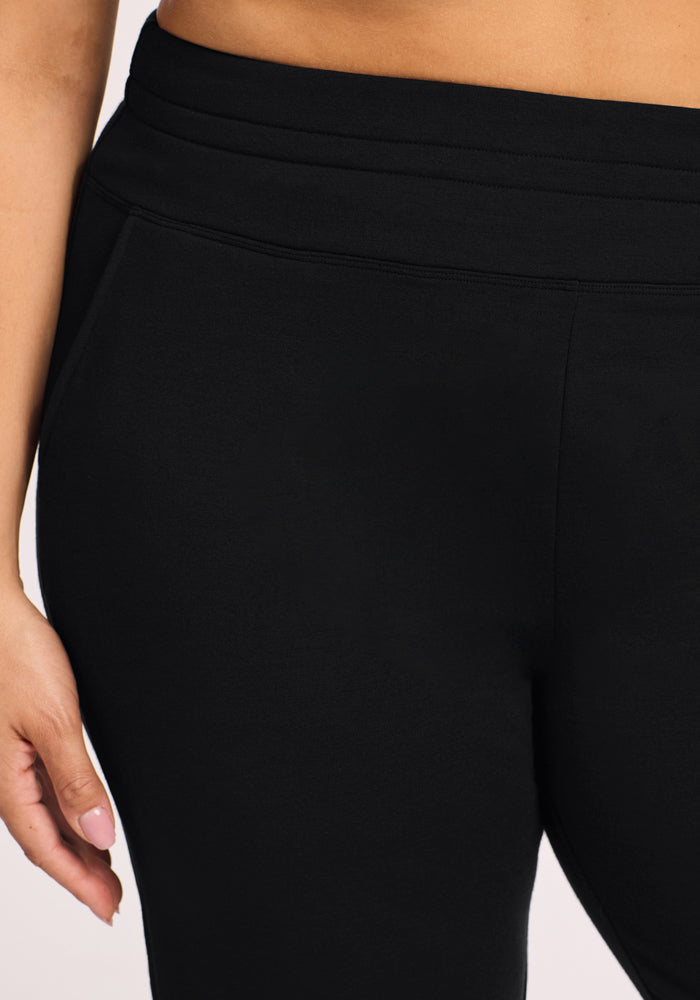 Close-up of a person wearing Woolx Willa Joggers in black, showing the waist, hips, and left hand with manicured nails. The plain light background highlights these comfortable joggers, perfect for pairing with lounge pants or merino styles.