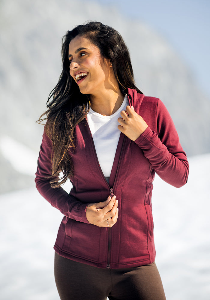 A smiling woman stands outdoors in snowy mountains, wearing the Woolx Zoey Zip Up Hoodie in Toasted Garnet over a white shirt and dark pants, enjoying the clear, sunny sky with one hand on the collar and the other on the zipper.